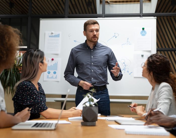 Confident businessman executive mentor ceo leader talk to employees group during corporate briefing in boardroom, serious male boss explain work plan to multiethnic workers at office meeting table